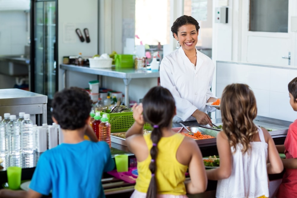 Vergabe Schulessen - Schüler und Schülerinnen stehen mit ihren Tabletts an der Ausgabe und warten auf Essen.